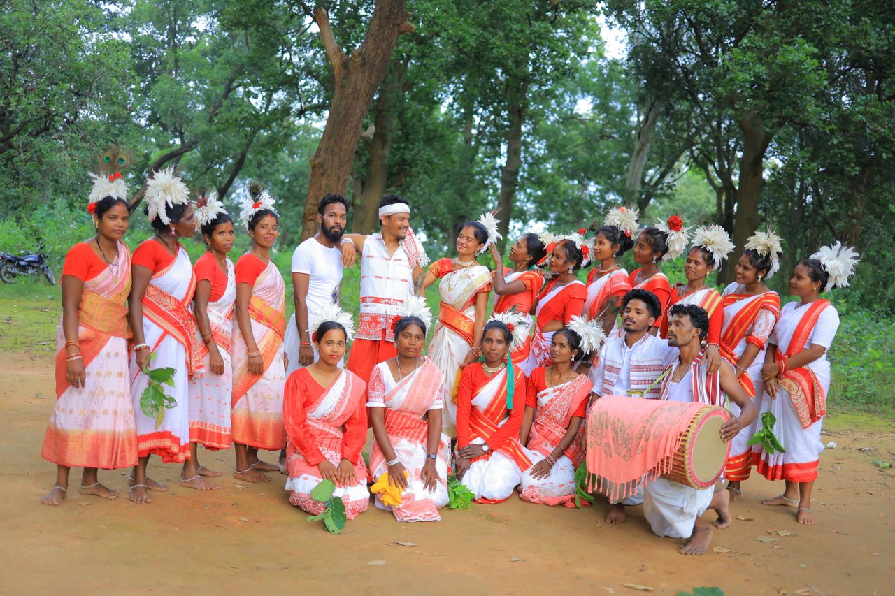 Group of adults in traditional red and white attire performing a tribal dance in a forest setting.
